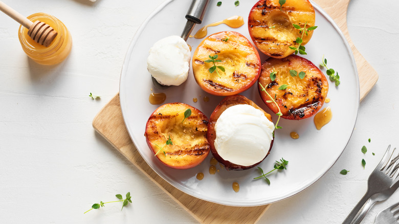 Grilled peaches decorated with thyme and a scoop of ice cream on a white plate next to a bowl of honey