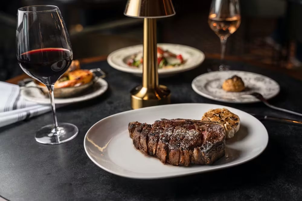 glass of red wine paired with a steak and garlic on a table next to a lamp with another glass of rose
