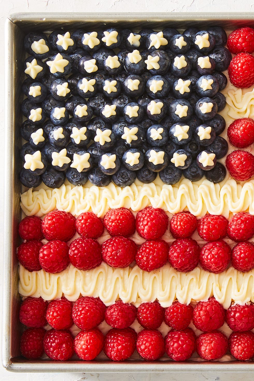 white cake decorated to look like an american flag with blueberries for stars, raspberries for red stripes, and off white frosting for white stripes