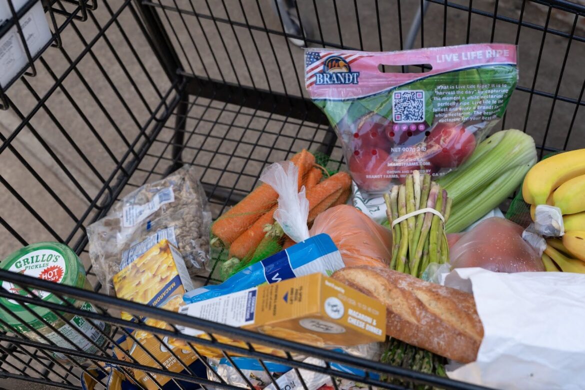 The Stakes: Struggling area food banks brace for higher demand after SNAP cuts A shopping cart full of food including bananas, carrots, apples, asparagus, long Italian bread, celery and boxes of pasta.