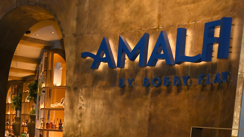 The sign for the Amalfi by Bobby Flay restaurant hanging on a brushed metal wall with a beautiful arch
