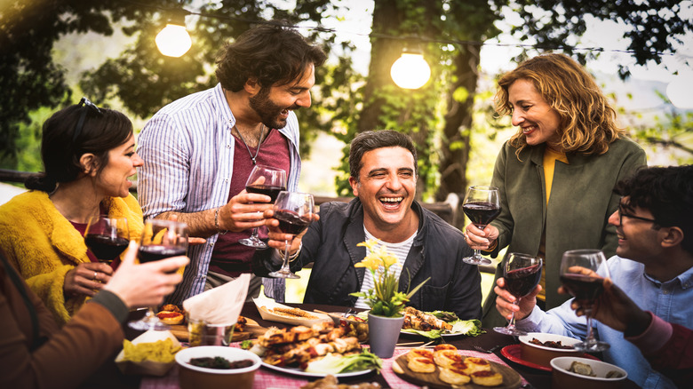 A group of men and women enjoying a bbq outside with food on the table and glasses of wine