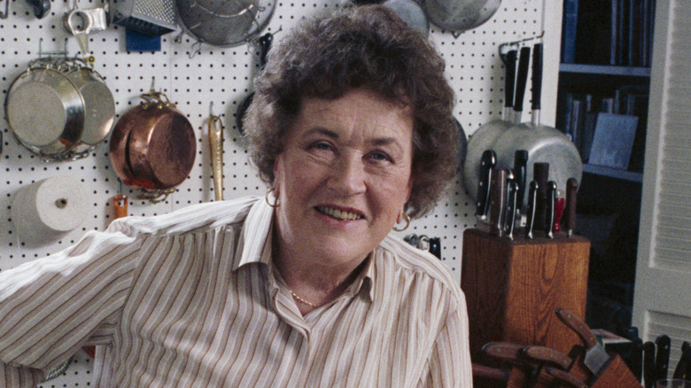 Julia Child smiling in front of wall with kitchenware hanging on it