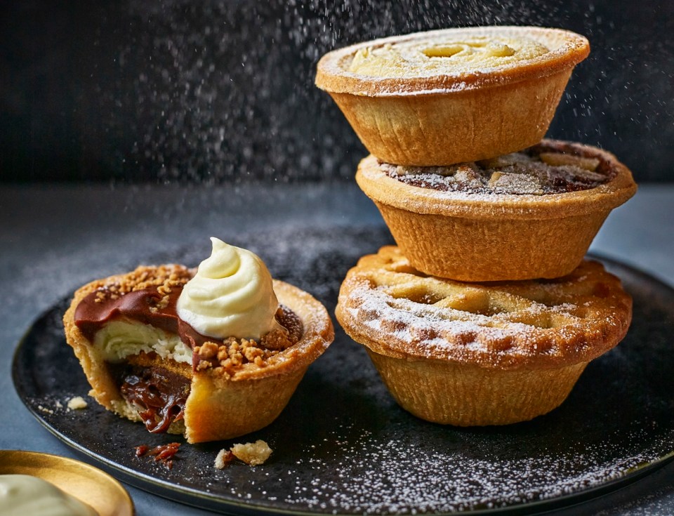 Three mince pies stacked on a plate, next to a partially eaten chocolate and cream-filled tart.