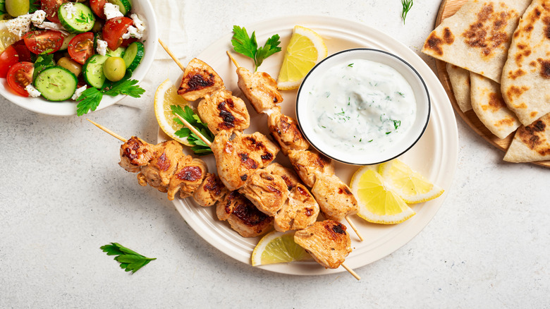 Chicken spiedini on a white plate with a yogurt sauces, lemons and a greek salad