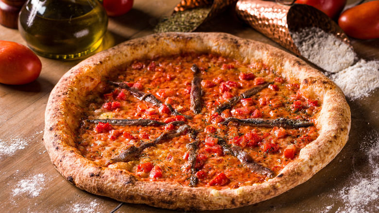 Anchovy pizza on a wooden counter with flour, herbs, olive oil, and a tomato