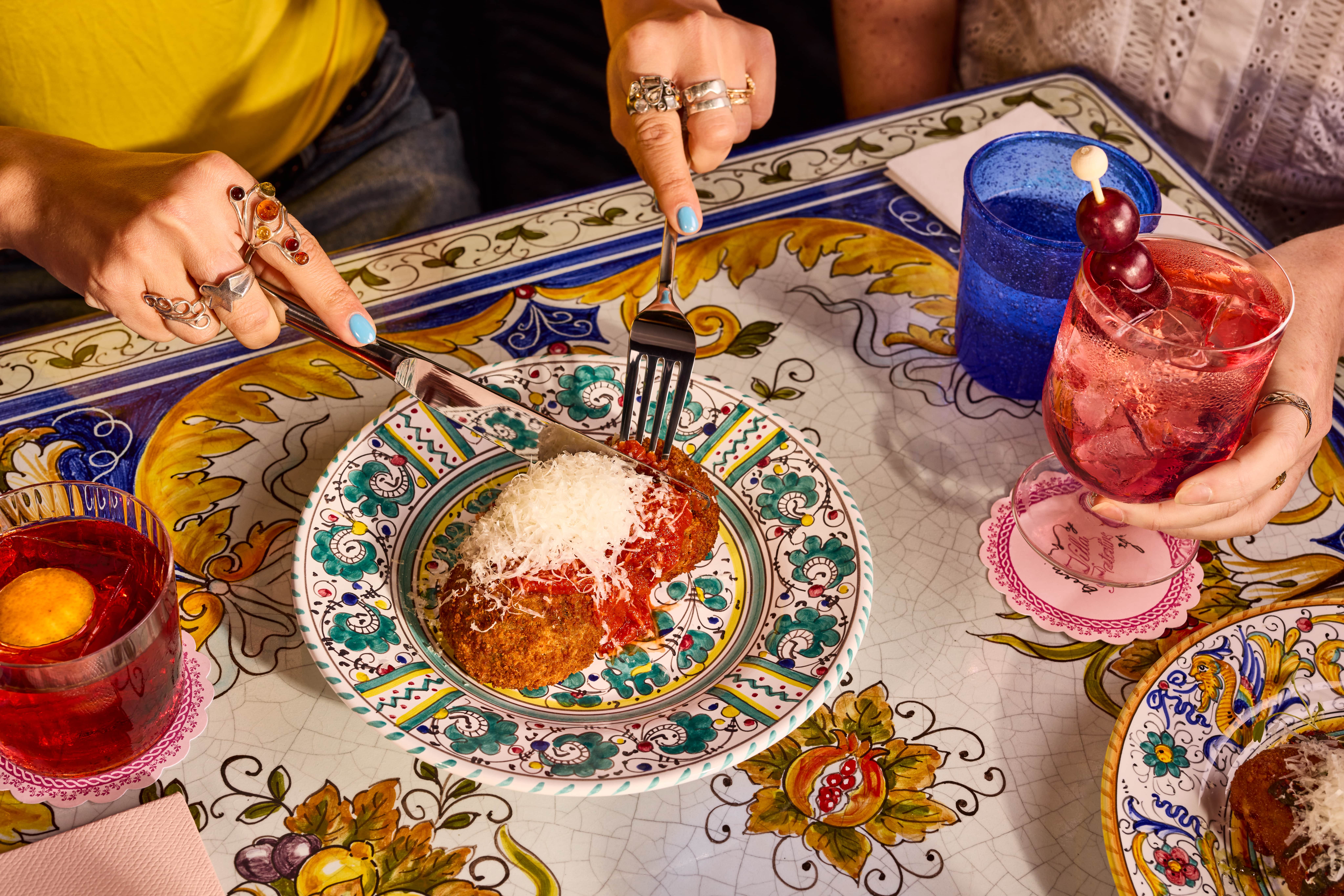 Two people eating breaded cutlets topped with tomato sauce and parmesan cheese at a table with colorful patterned plates and drinks.