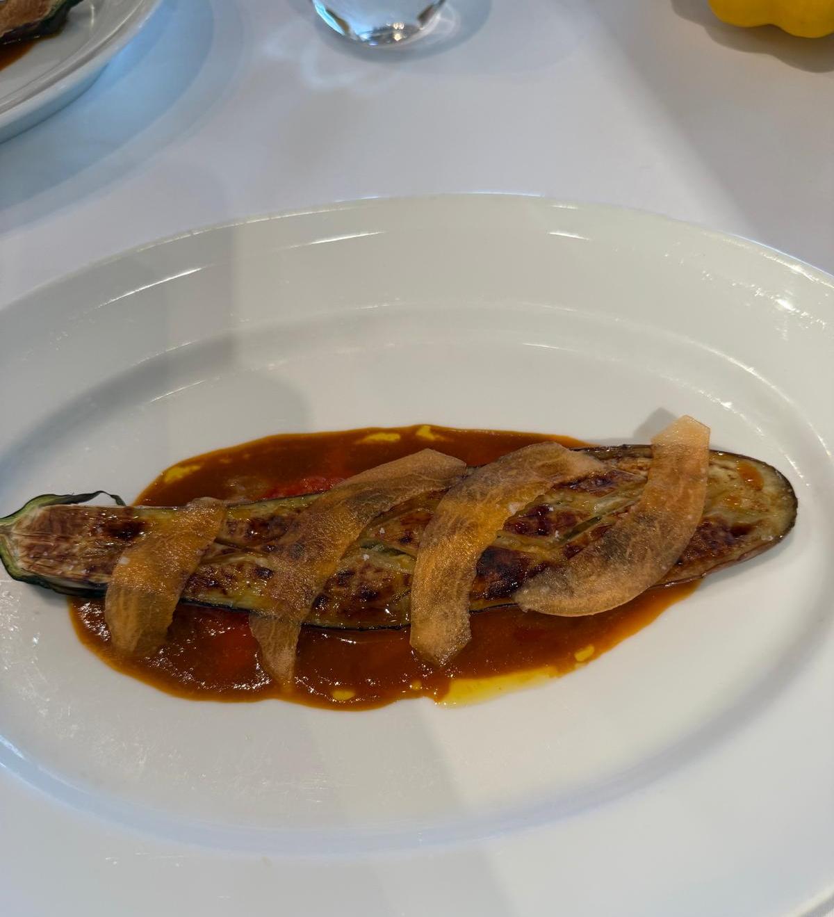 Roasted eggplant with sauce and crispy vegetable garnish on a white plate.