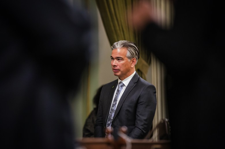 A person with short, slicked-back salt-and-pepper hair stands in a formal setting, wearing a dark suit, white shirt, and patterned tie. Green curtains hang in the background, and blurred figures in dark clothing frame the foreground.