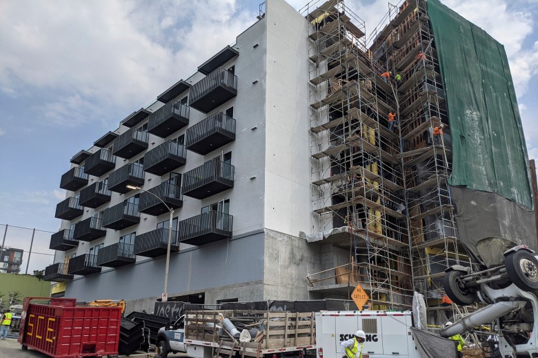 Construction workers are working on scaffolding on a five-story apartment building on a sunny day. On the ground floor, trucks and other construction tools can be seen parked near the building, and a construction worker is walking nearby in a hard hat.