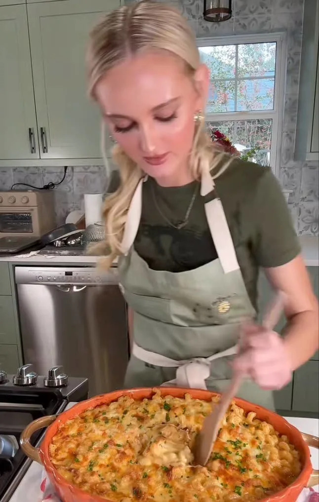 Person stirring a baked pasta dish in a kitchen, wearing a casual shirt and apron