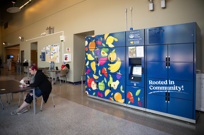 A colorful food locker station labeled “Rooted in Community!” stands against a wall inside a Contra Costa College building. The locker, decorated with illustrations of fruit, is a partnership with the Food Bank of Contra Costa and Solano. A person with a knee brace sits at a nearby table eating, while another person uses a laptop against the wall. Bulletin boards and hallway doors are visible in the background.