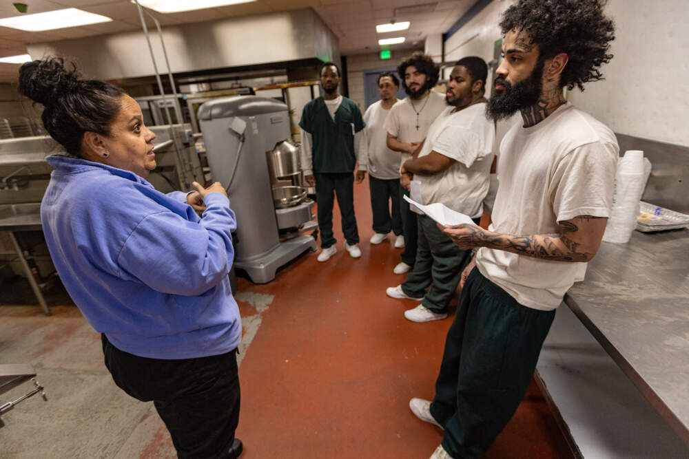 Trainer Rebecca Queeney talks with class participant Marino Pimentel as the other students listen. Queeney teaches the men everything from proper chopping techniques to how to make common sauces, entrees and desserts. (Jesse Costa/WBUR)