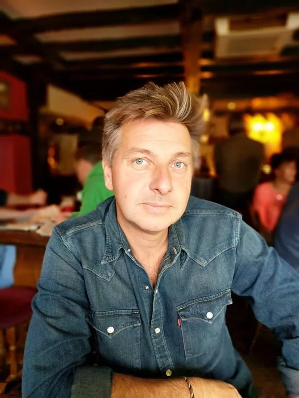 A recent portrait shot of wine expert Andy Pincott sitting at a table wearing a denim shirt