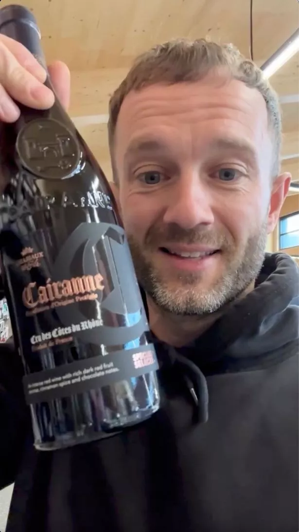 A picture of Liam D'Arcy, wearing a black hoodie, and holding up a bottle of wine