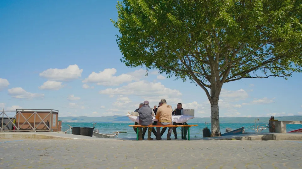Stanley Tucci and local fisherman eat at a picnic table alongside a lake in Tucci in Italy