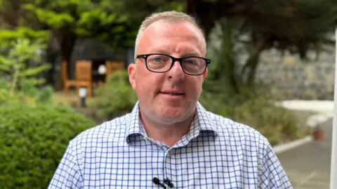 A headshot of a man with short brown hair and black glasses. He is wearing a black and blue checked shirt.