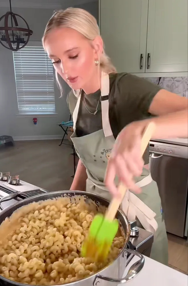 Person cooking pasta in a kitchen, wearing an apron and stirring with a spatula over a stove