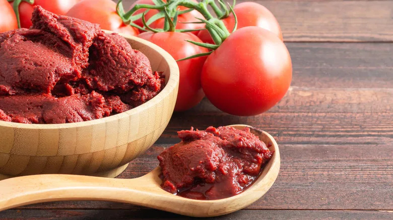 A large amount of tomato paste in a wooden bowl sits beside ripe tomatoes and a wooden spoon full of tomato paste