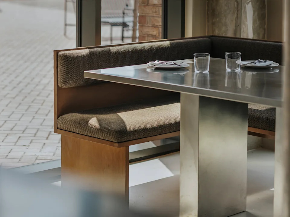 A restaurant with brutalist interiors features wooden upholstered banquettes with warm, brown fabric matching with matte chrome tables with a geometric shape.