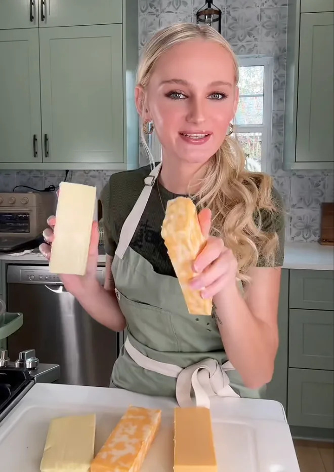Person in a kitchen holding cheese blocks, wearing an apron over a green top