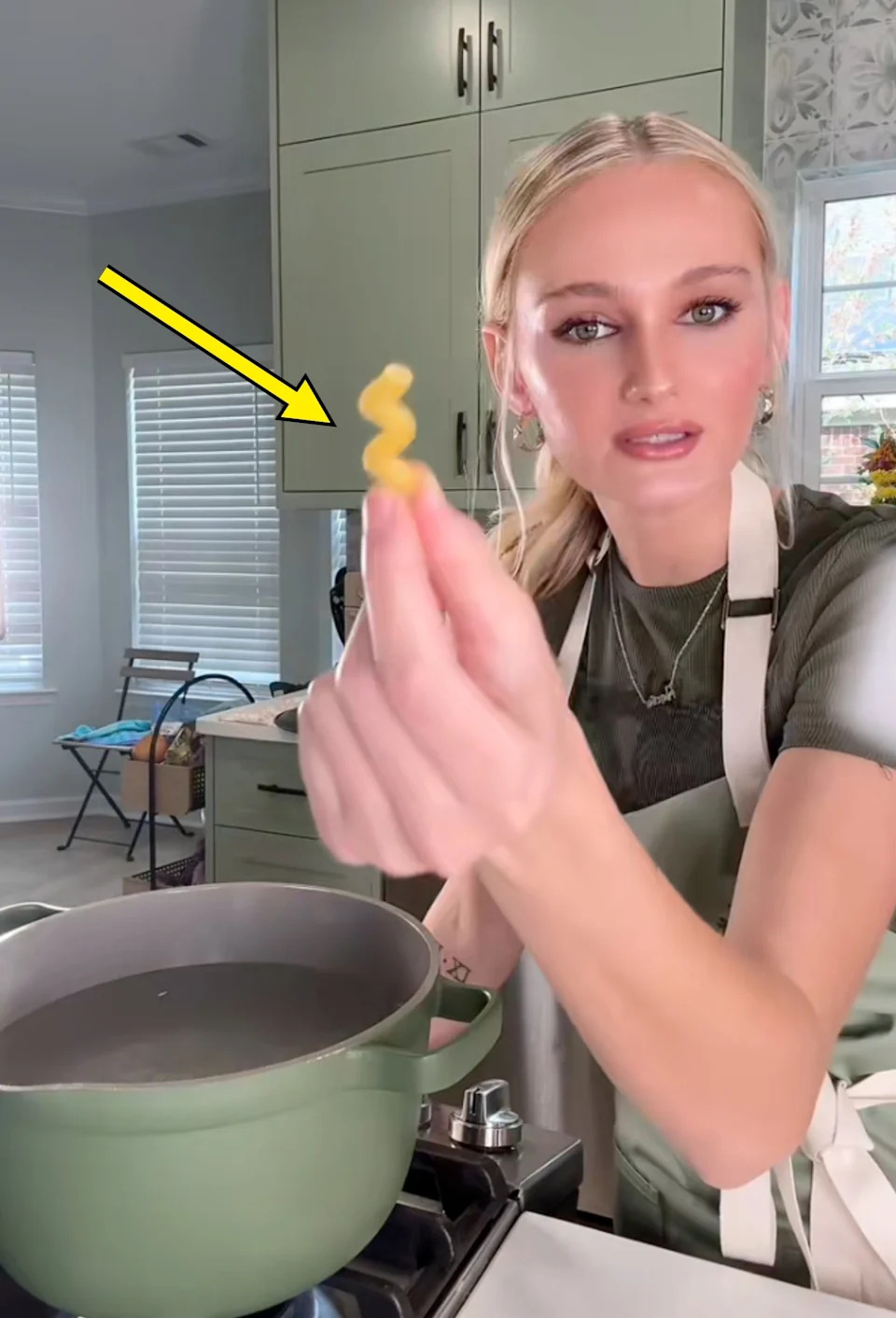 Person holding a piece of pasta above a pot on a stove, wearing a casual shirt and apron, in a kitchen setting