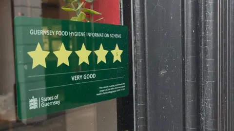 BBC A photo of a green sticker on a window. The sticker has five stars on it and is titled 'Guernsey Food Hygiene Information Scheme'