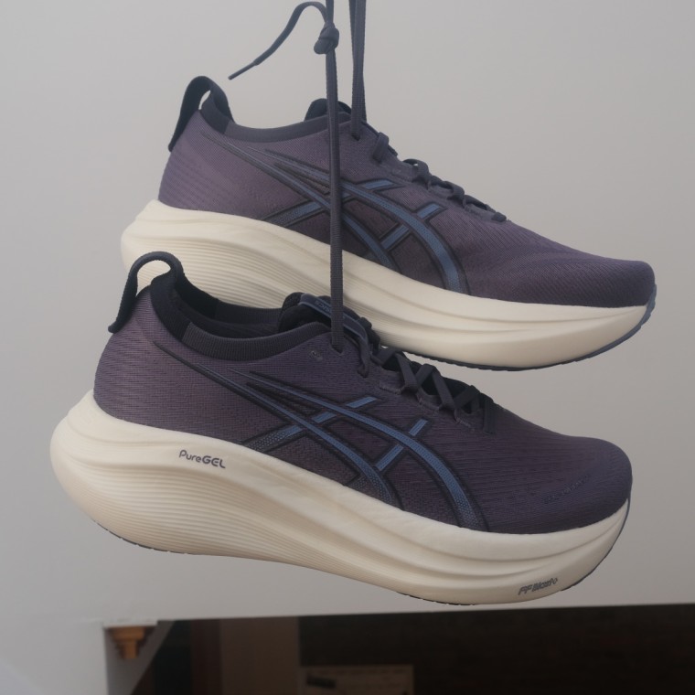 A close up shot of the Asics Gel-Nimbus 27 in the purple colorway, hanging by the laces, against a white background.