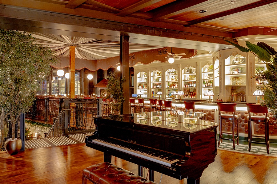 Interior of a restaurant features a grand piano, a bar upholstered in brown leather with a white wooden backbar, and a pale green-and-white-striped canopy over the dining room.