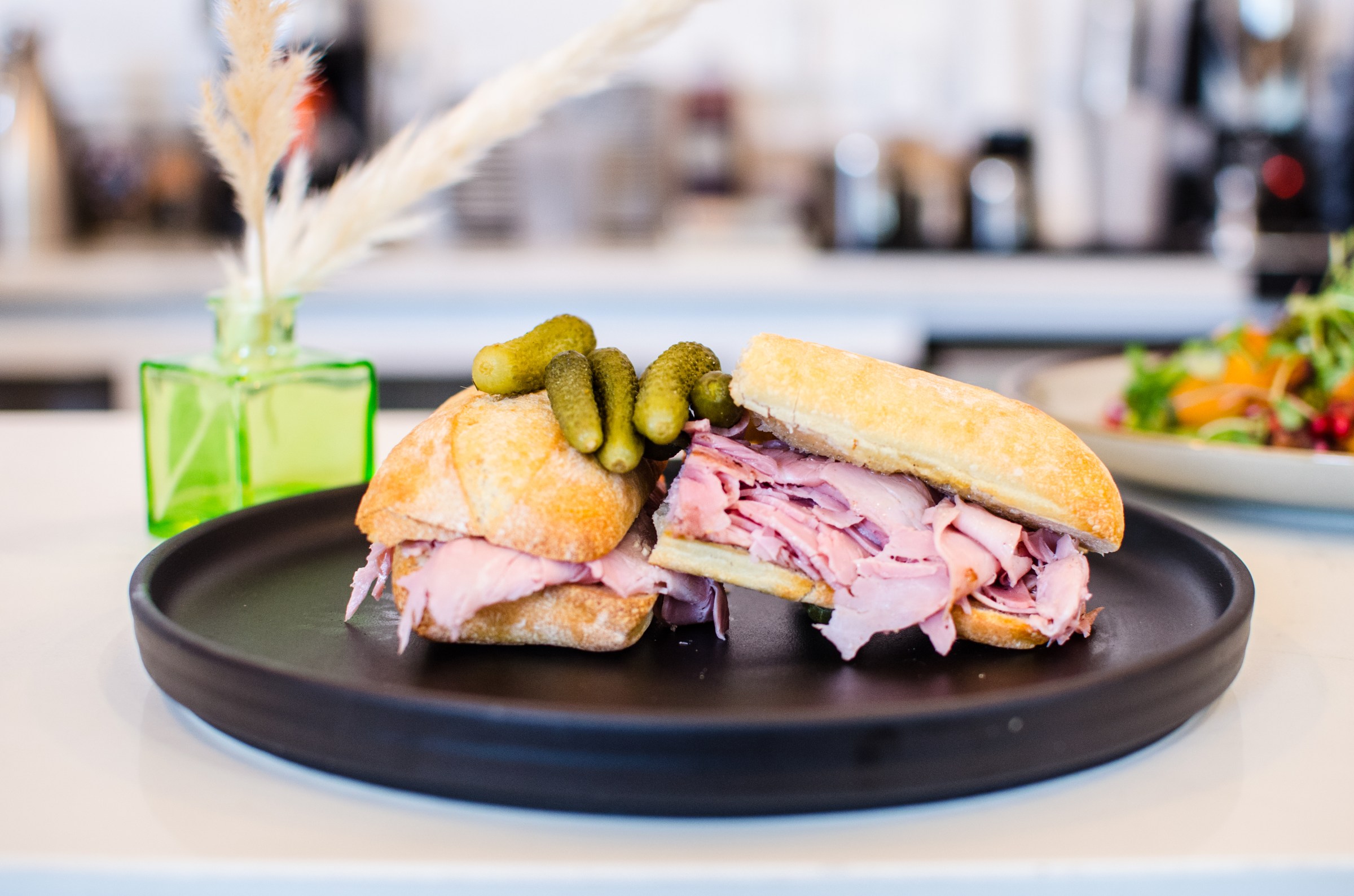 Thin slices of ham are sandwiched on a baguette, topped with mini pickles. It all sits on a black plate on a white counter.