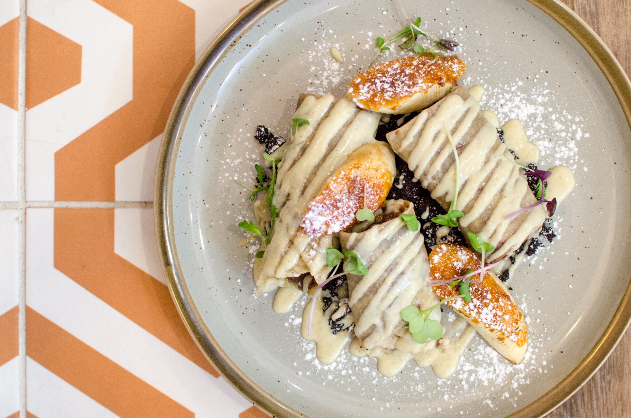 Overhead view of a restaurant dish. A rolled up crepe is sliced in thirds, drizzled with a cream sauce, and topped with bruleed banana slices. 