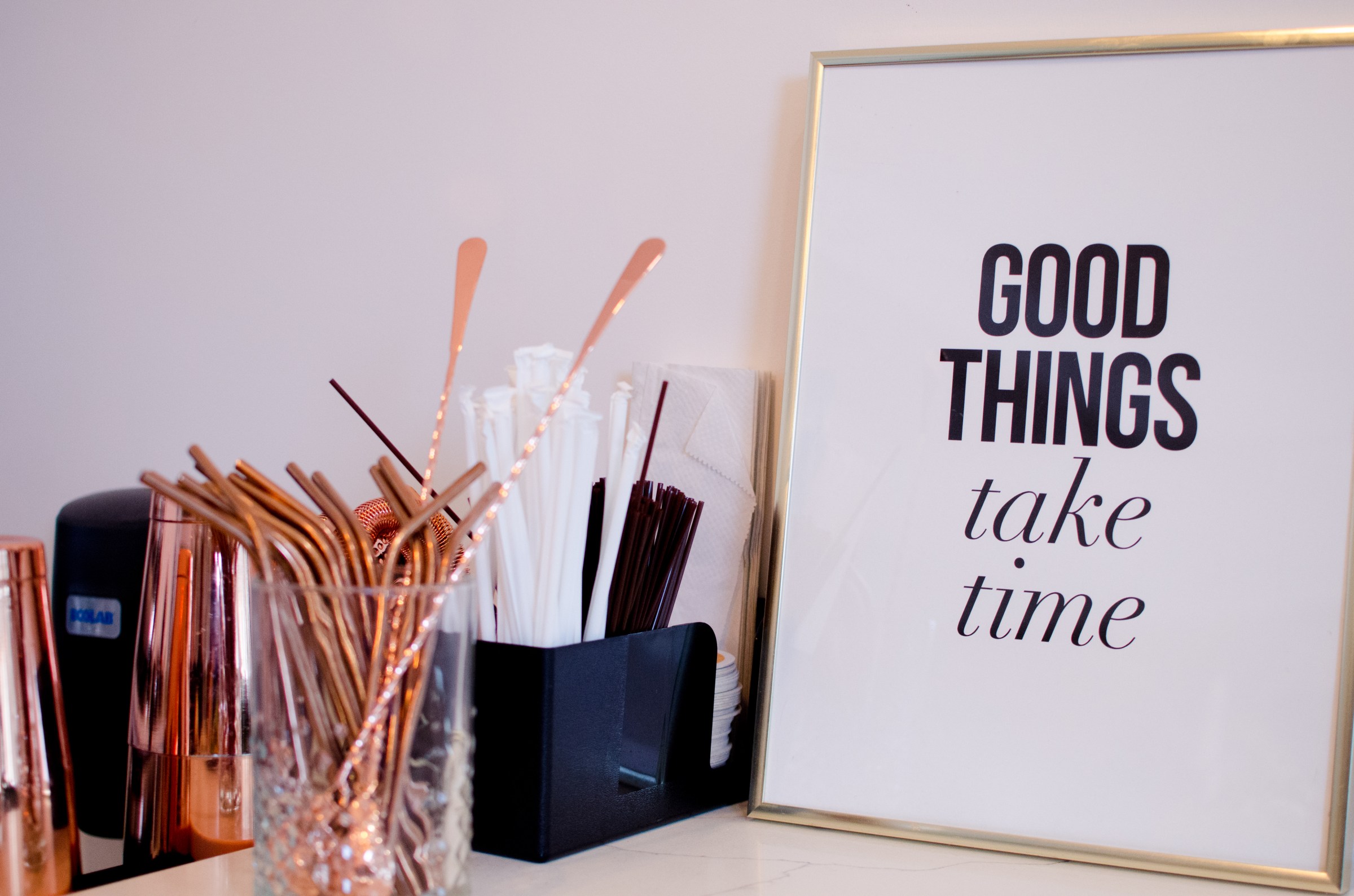 A white sign that says “Good things take time” leans against a wall. A jar of metal straws and other beverage accessories are near.