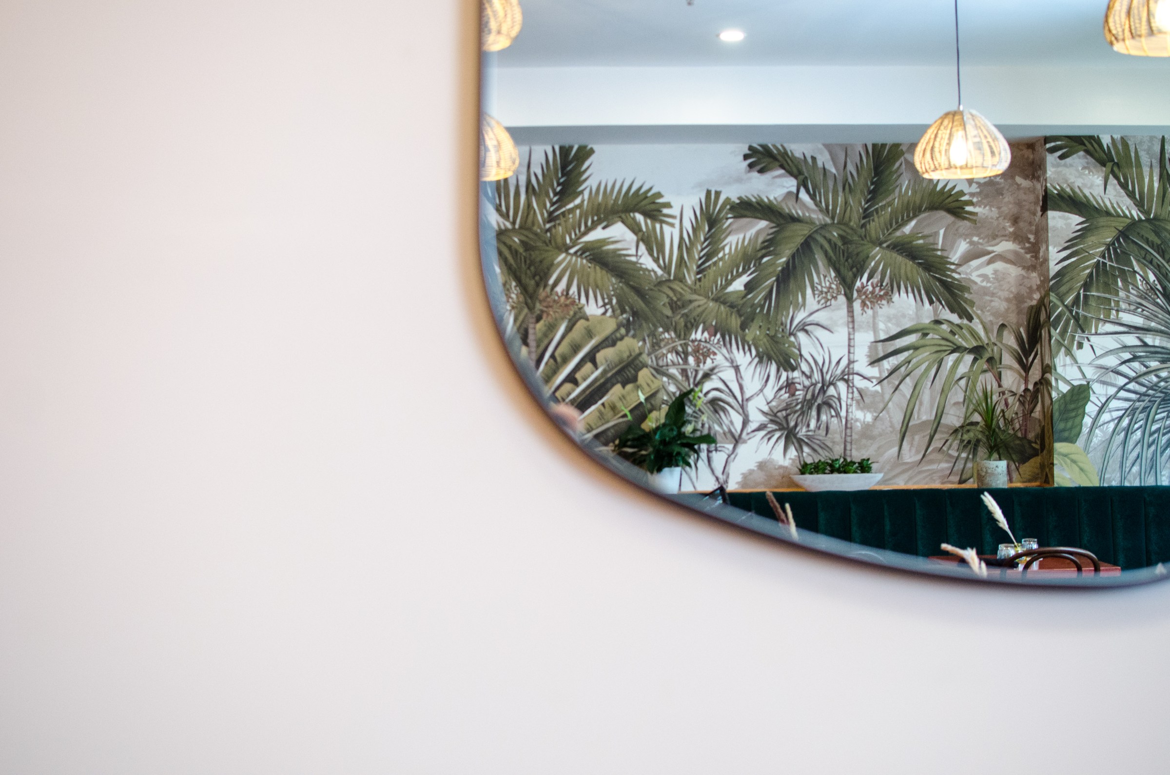 On a white wall, a mirror with a rounded edge takes up nearly half the photo, showing a vintage jungle wallpaper-covered wall opposite