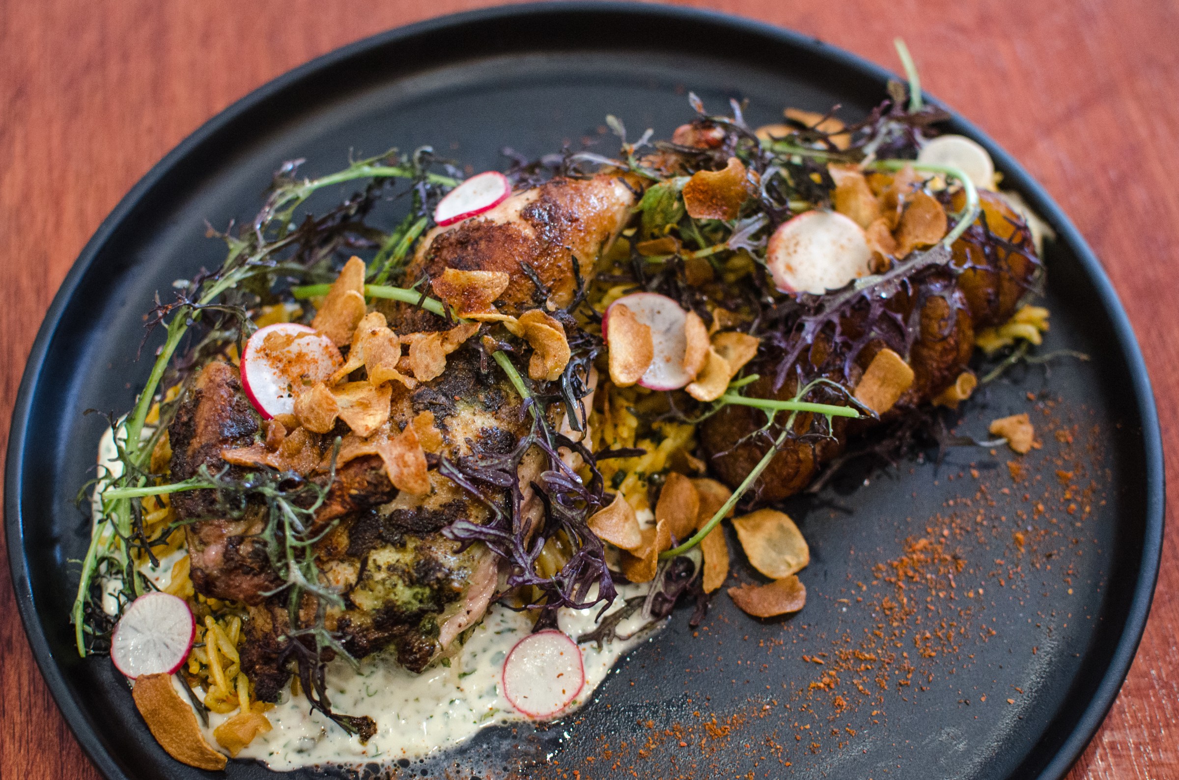 A matte black plate holds a roast chicken covered with crispy garlic, sliced radishes, and greens. A red spice dots the plate.