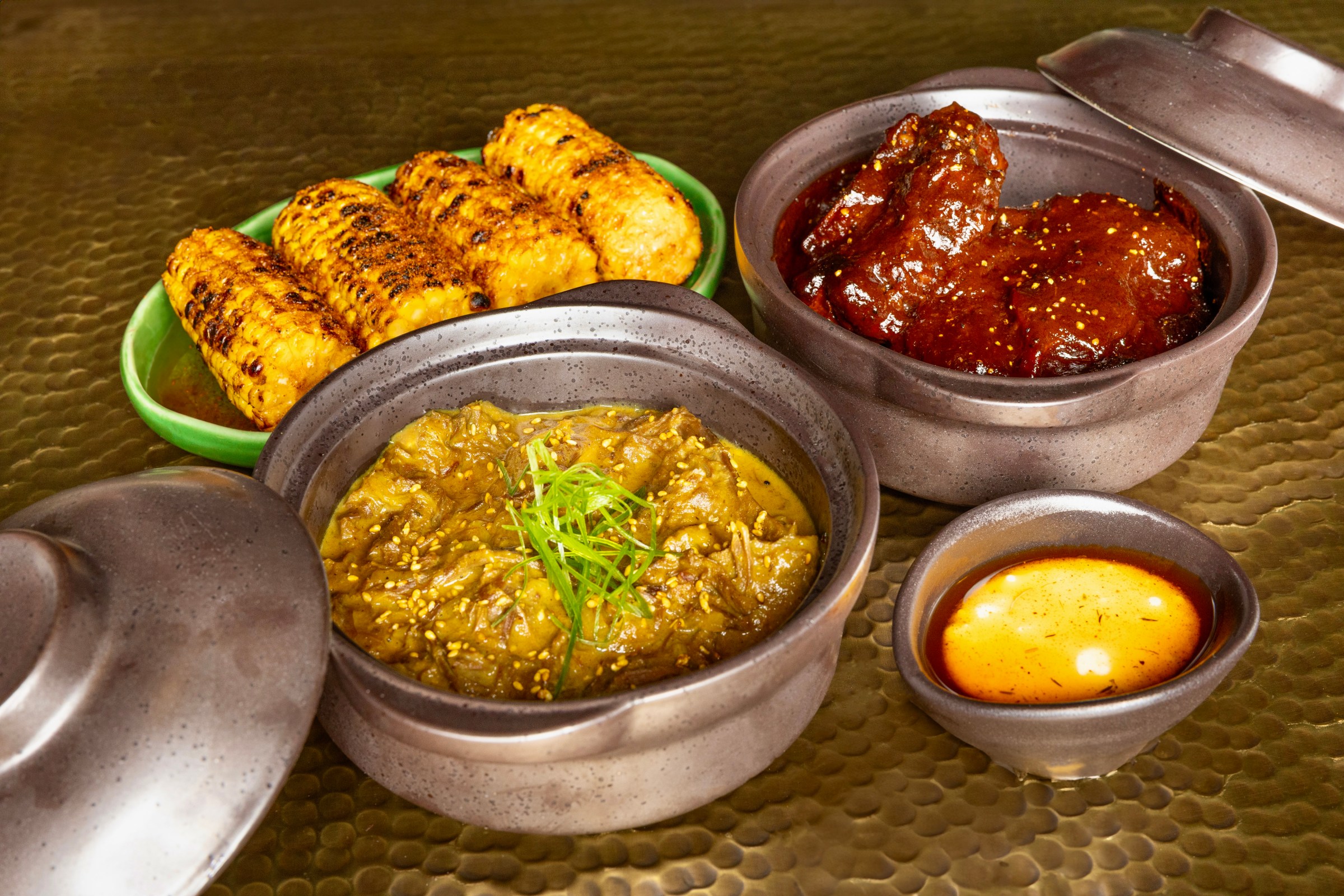A table of a plate of grilled corn, another pot of curry, and a third with brown sauce-covered meat.