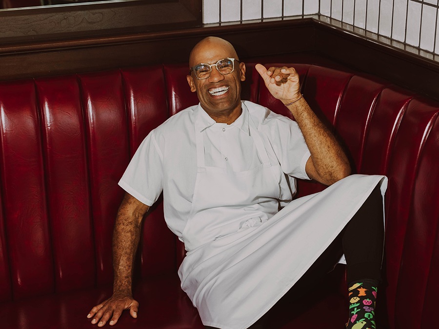 A smiling man, wearing glasses and a white shirt and apron, sits relaxed on a red leather banquette.