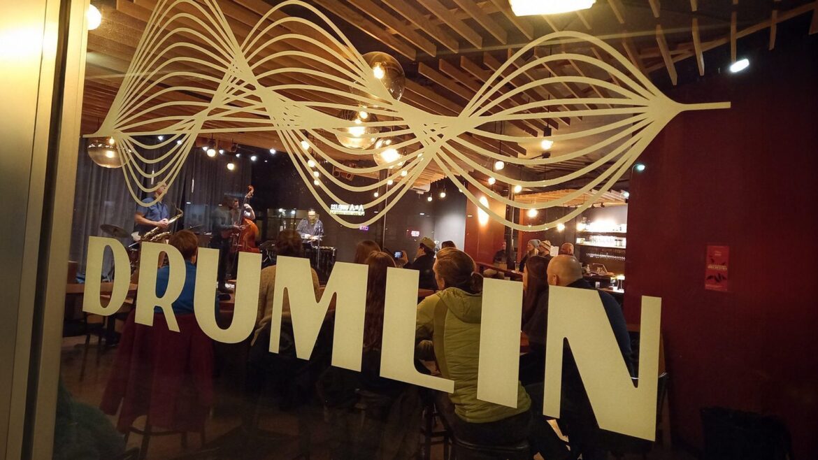 Peering through the window marked "Drumlin," the dimly lit interior reveals a space that has reinvented itself, with people seated and a band playing evocatively on stage.