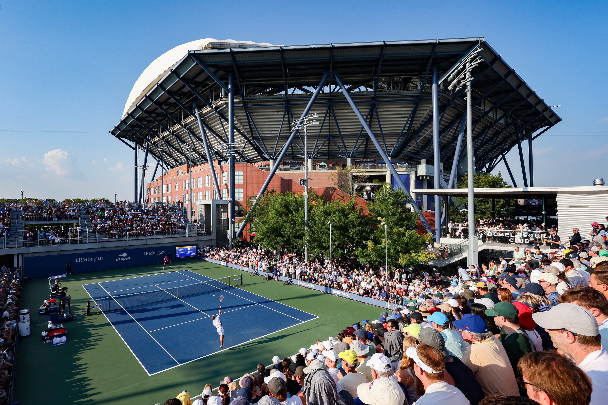 An outdoor tennis match with a tennis stadium in the background.