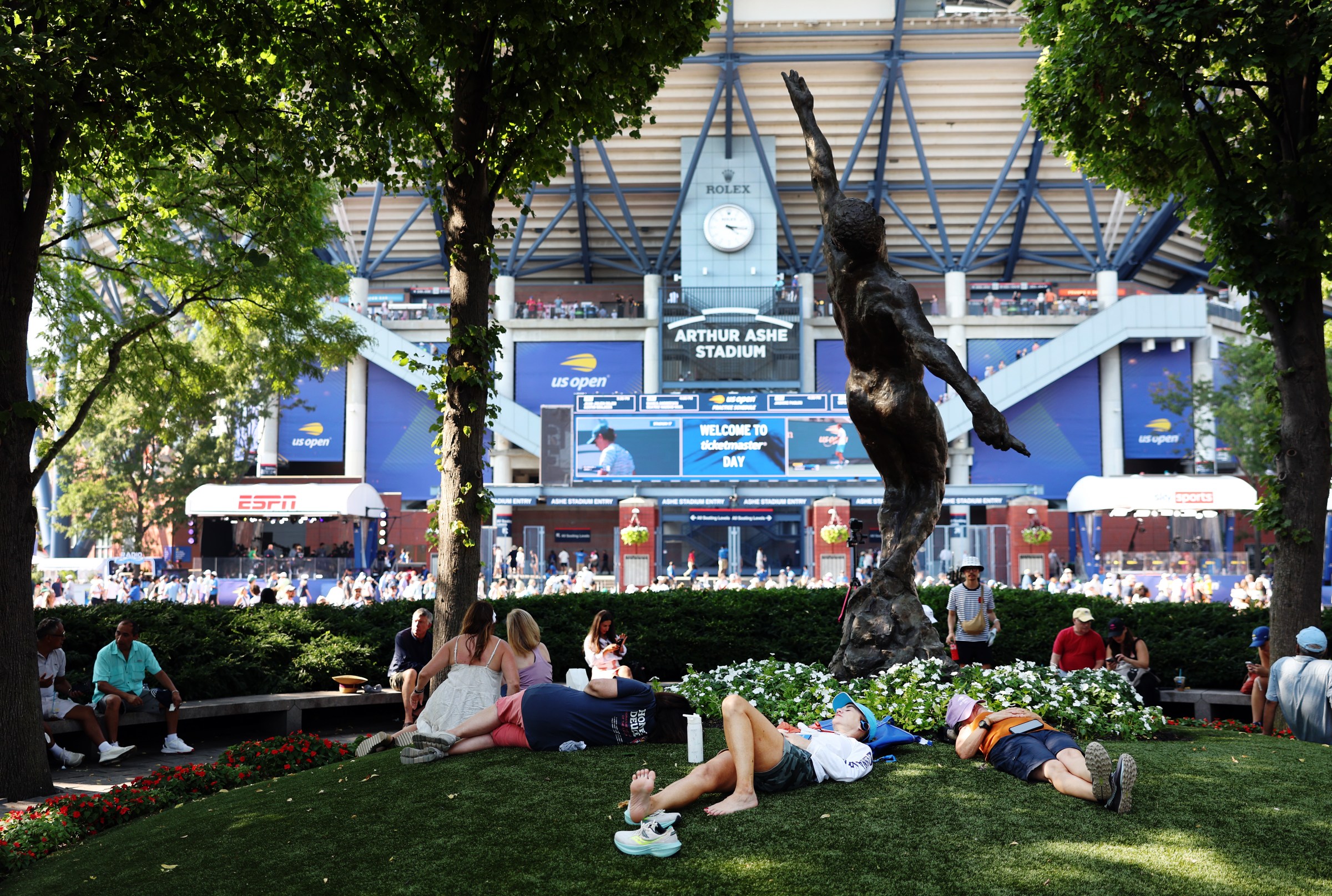 People laying down on grass underneath a statue in front of a stadium.