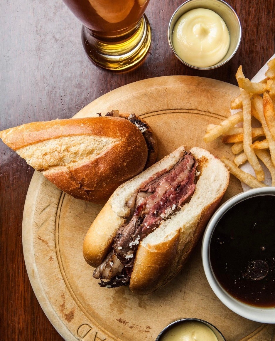 An angled-halved meat sandwich on a round wooden plate with a cup of dark brown broth.