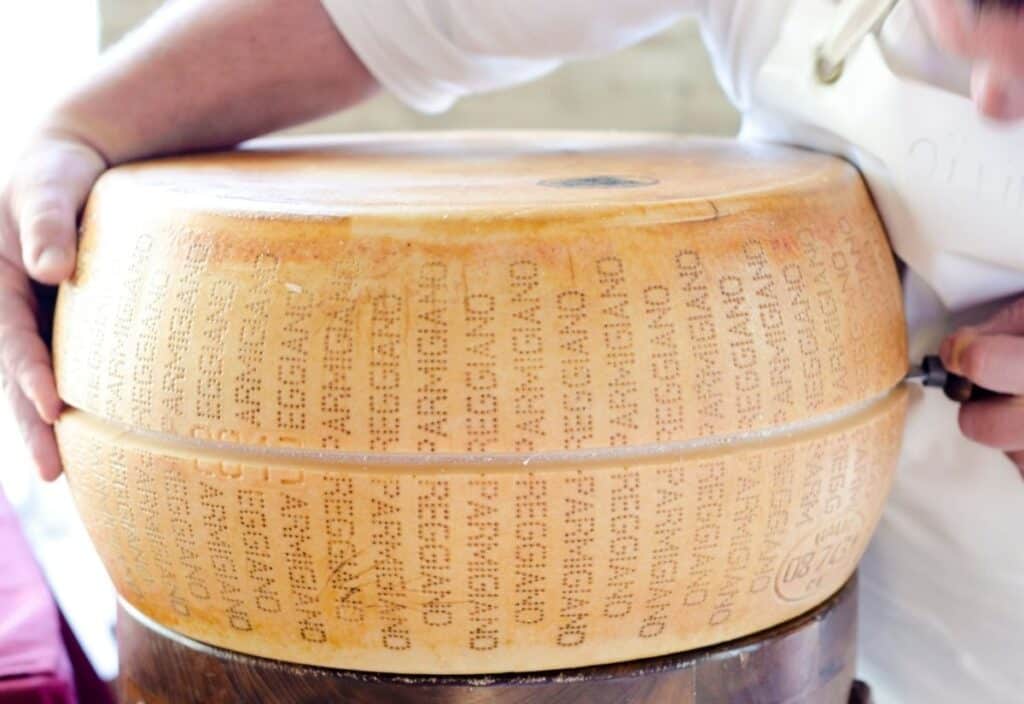 A person cuts a large wheel of Parmigiano Reggiano cheese, stamped with its name on the rind.