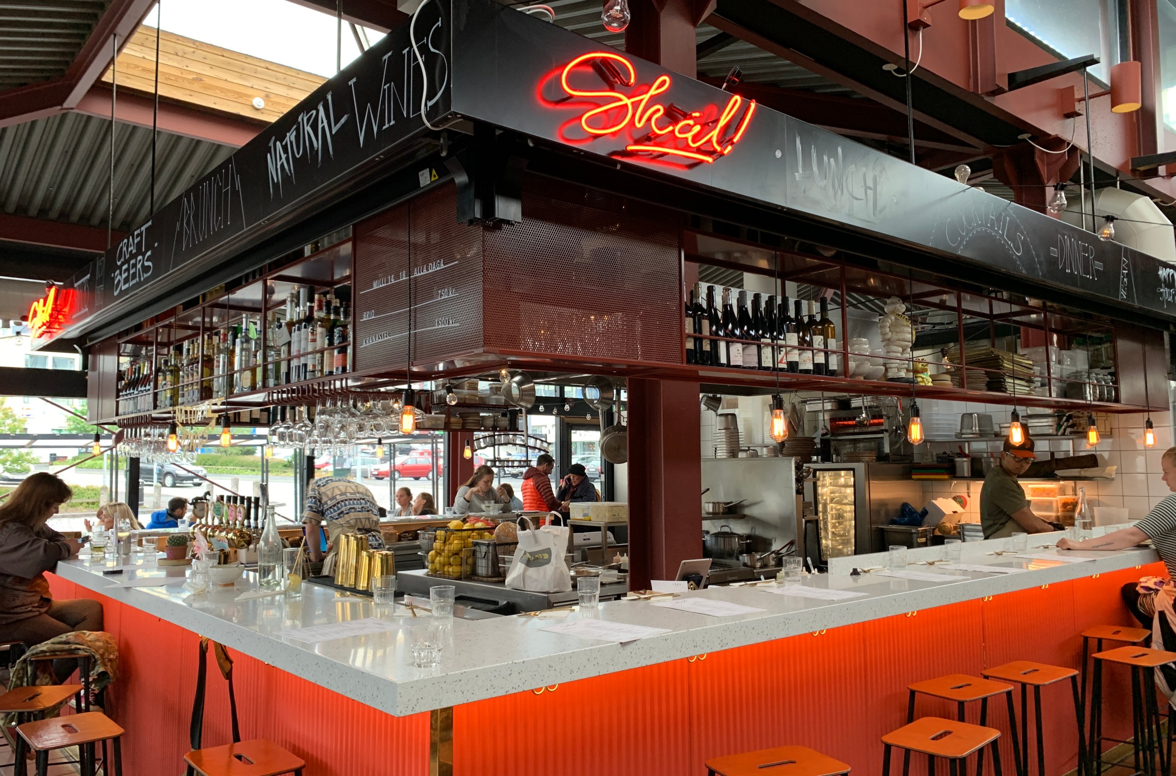 A large U-shaped counter surrounds a bar and open kitchen beneath a bright neon sign inside an industrial warehouse space.