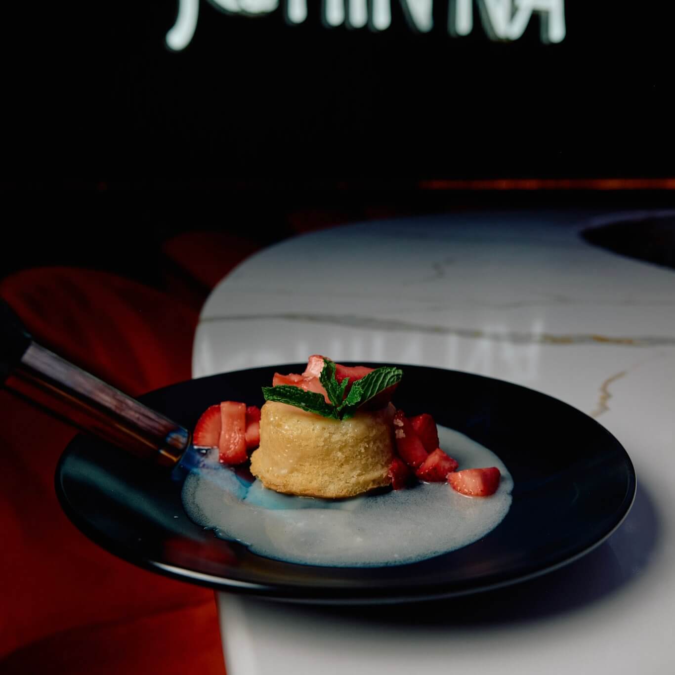 A dessert topped with mint and diced fruit sits on a dark plate with sauce, placed on a white marble table. A torch is visible on the left side.