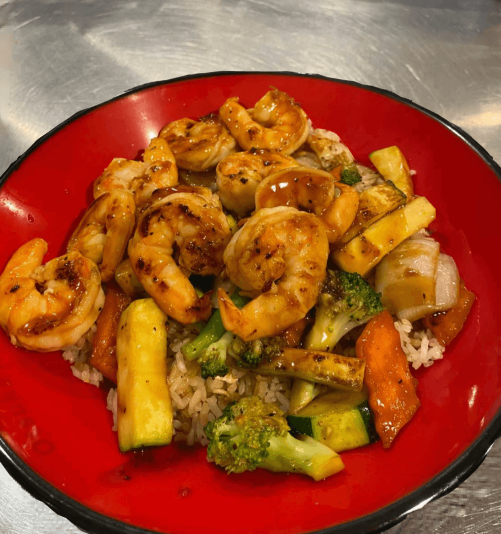 A red bowl filled with grilled shrimp, sautéed zucchini, carrots, broccoli, onions, and brown rice, topped with a glossy sauce.