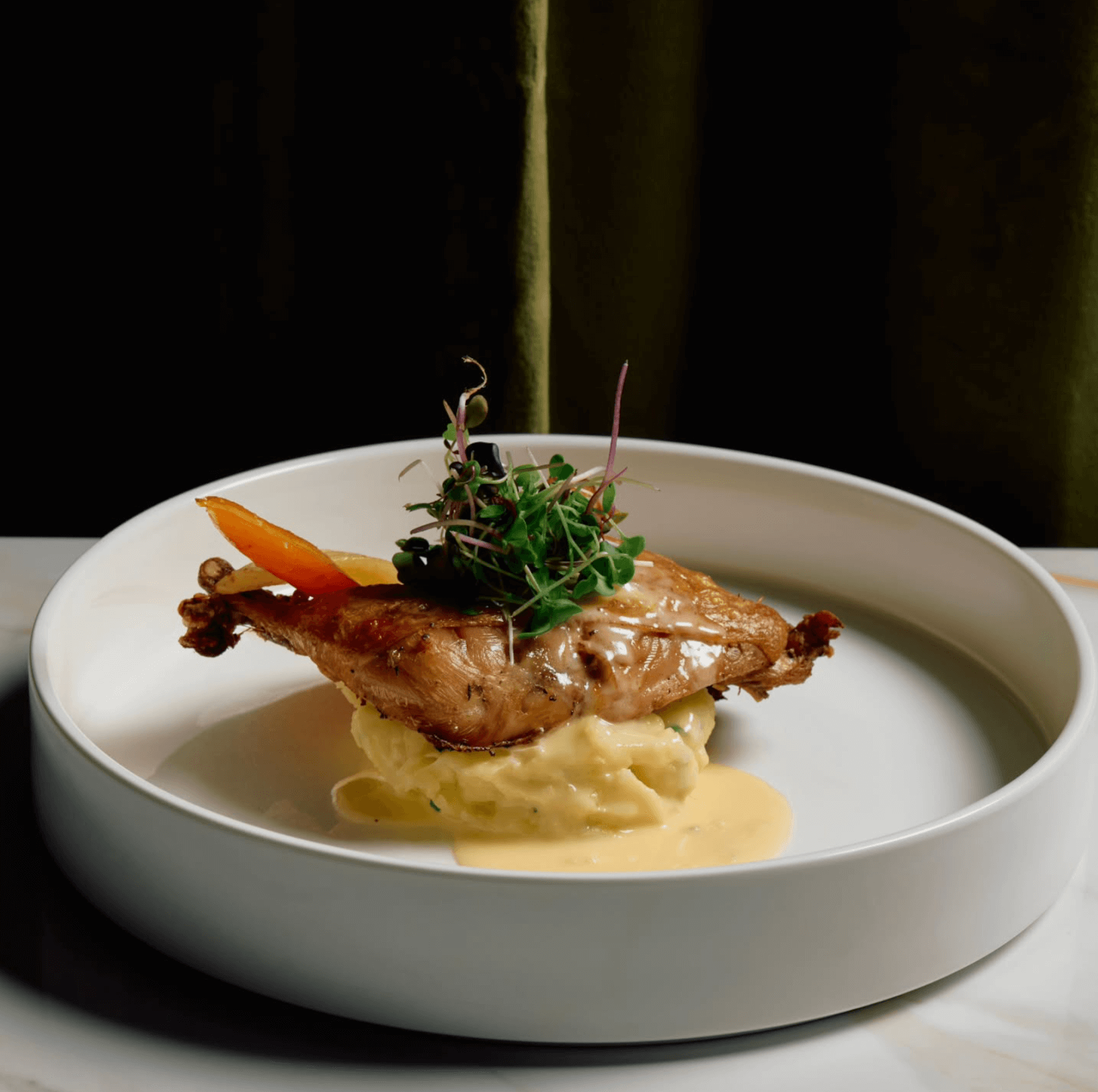 Roasted chicken thigh on mashed potatoes with creamy sauce, garnished with microgreens and a carrot slice, served in a white bowl—a highlight among Birmingham restaurants' new dining concepts.