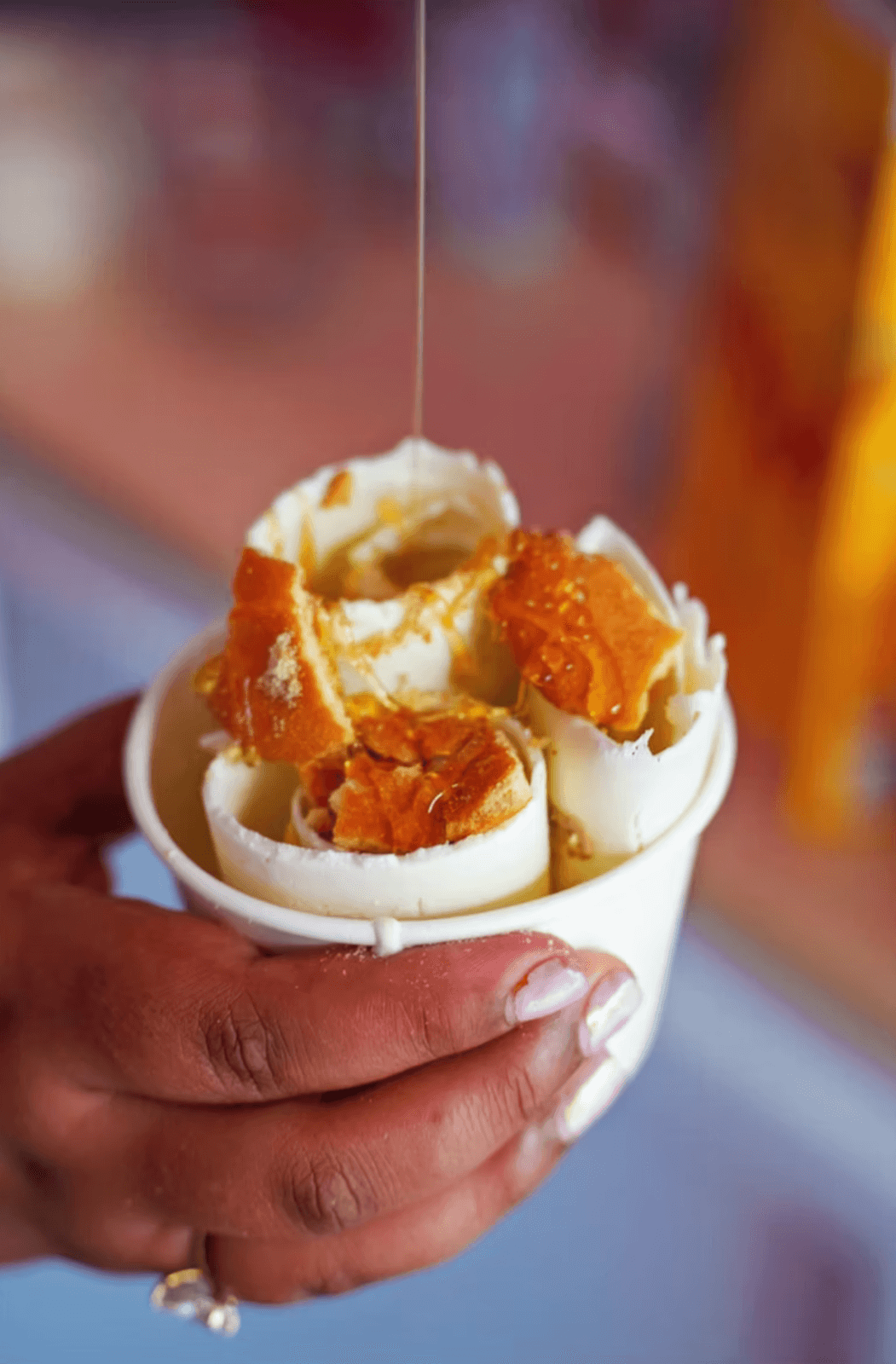 A hand holds a cup of rolled ice cream topped with honeycomb pieces, while syrup is being drizzled over it.