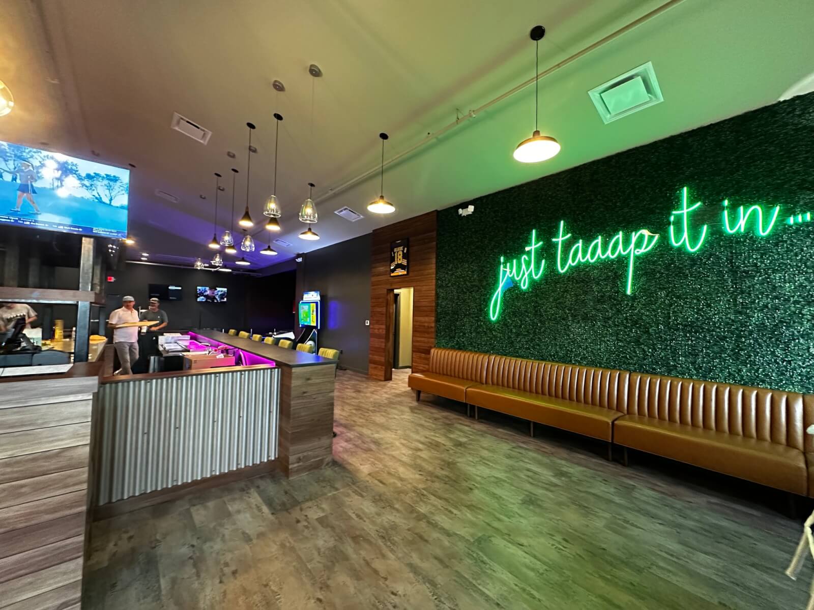Modern bar interior inspired by innovative restaurant concepts, featuring a neon sign reading "just taaap it in...", leather bench seating, hanging pendant lights, and a TV showing golf.
