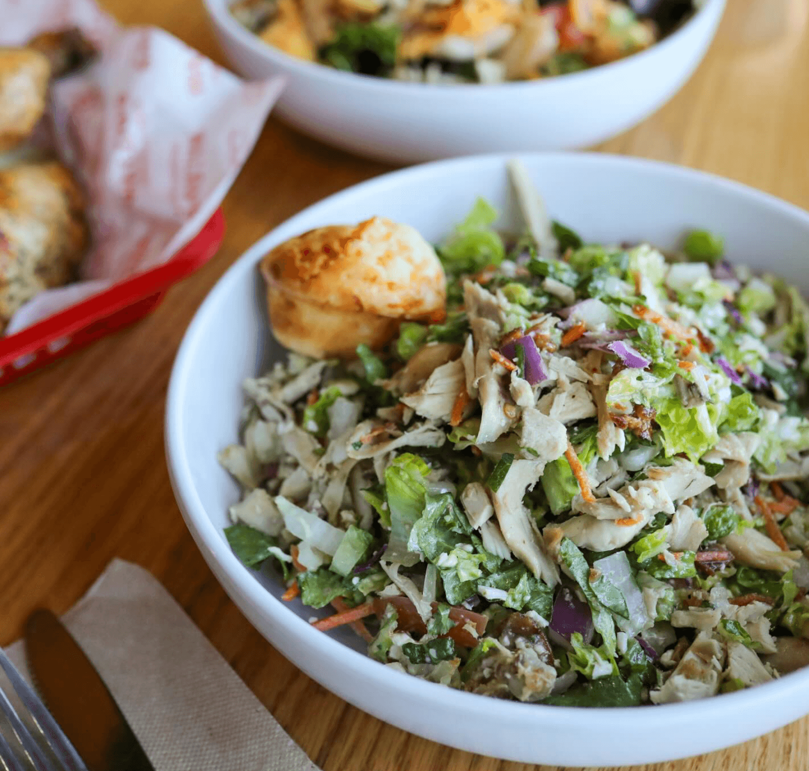A bowl of chopped chicken salad with mixed greens and vegetables, topped with a small biscuit, sits on a wooden table—showcasing one of Birmingham restaurants’ fresh dining concepts alongside another salad and baked goods.
