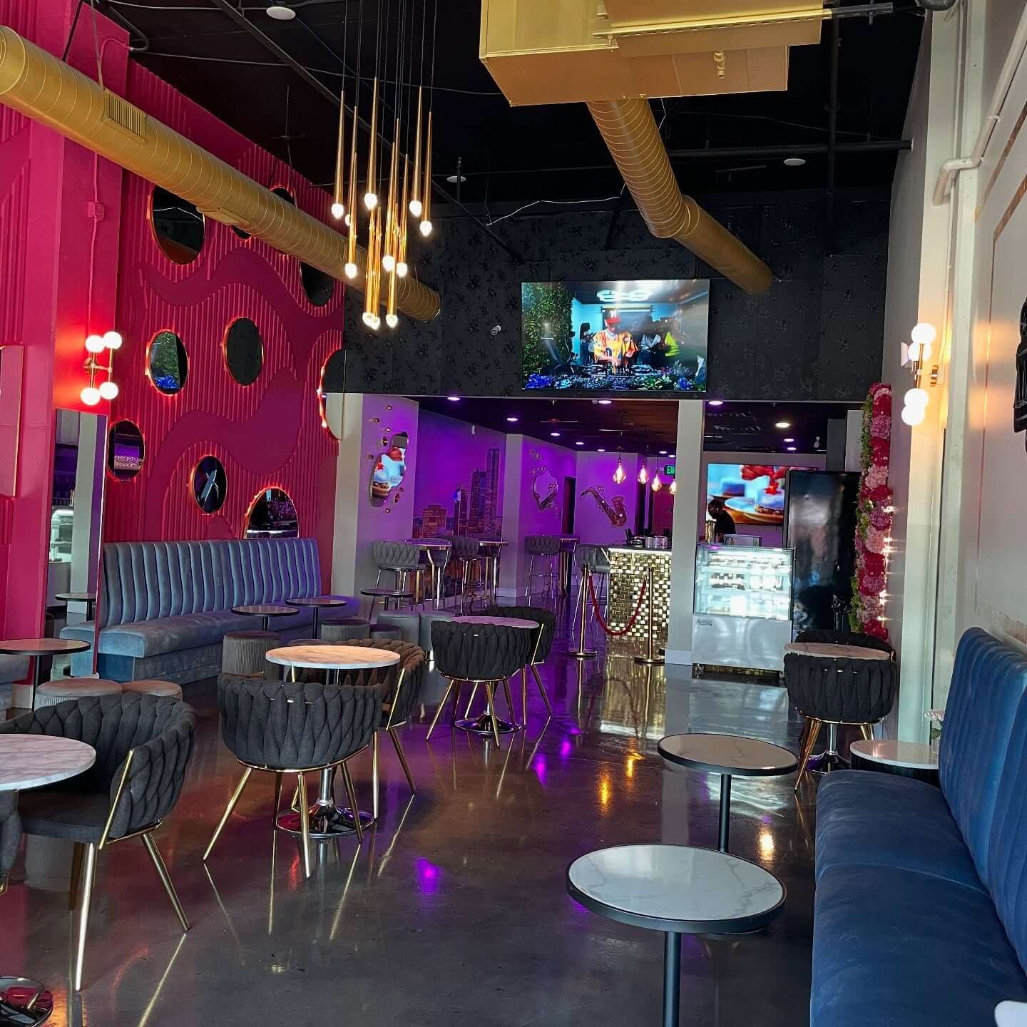 Modern lounge with colorful decor, round tables, blue and gray seating, pink accent wall, hanging lights, and a TV screen in the background—a stylish look for new dining concepts among Birmingham restaurants. The space appears empty.