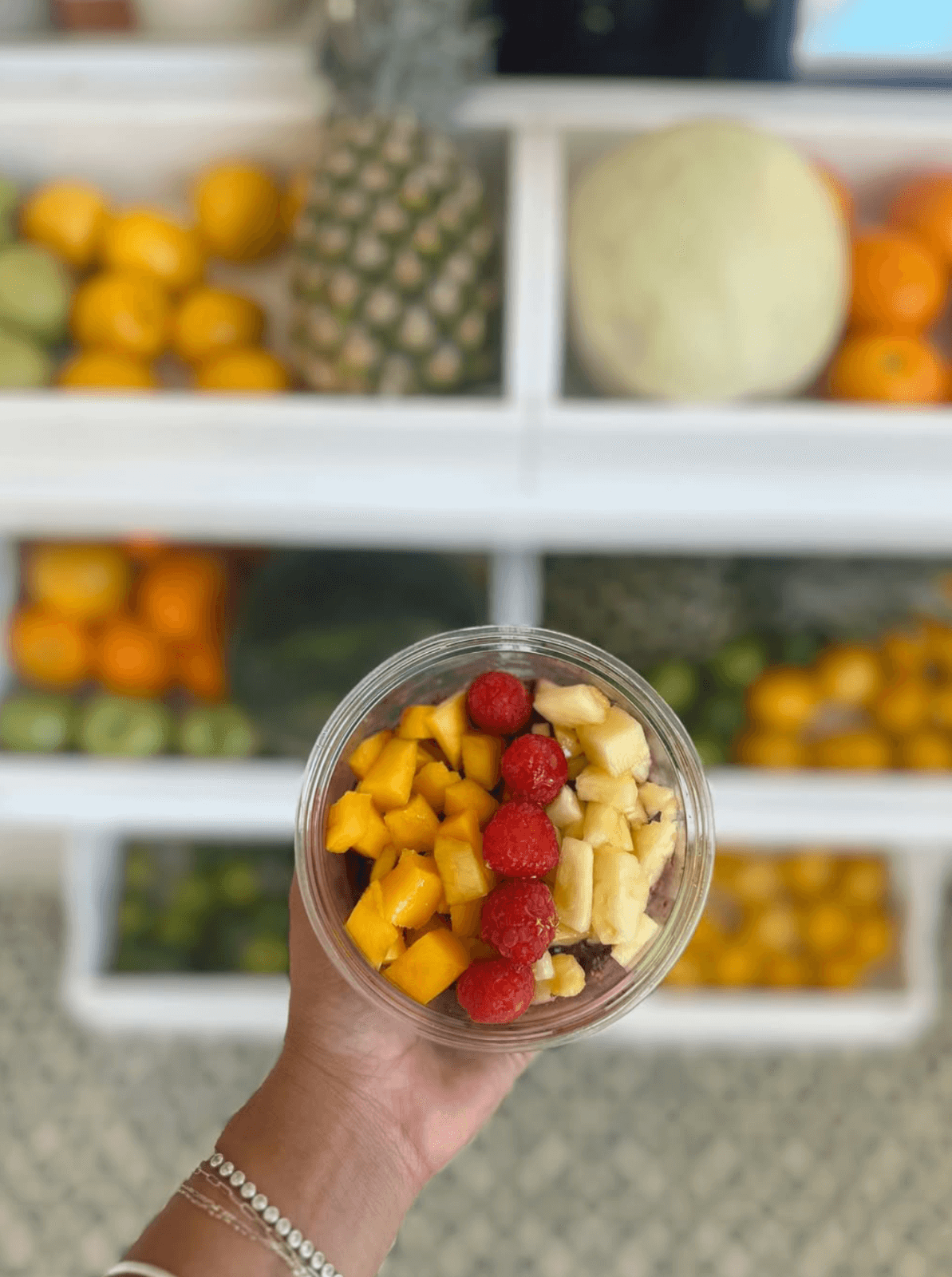 A hand holds a bowl of chopped fruit—mango, banana, and raspberries—in front of shelves filled with whole fruits, capturing fresh inspiration often found in new restaurants and innovative dining concepts.